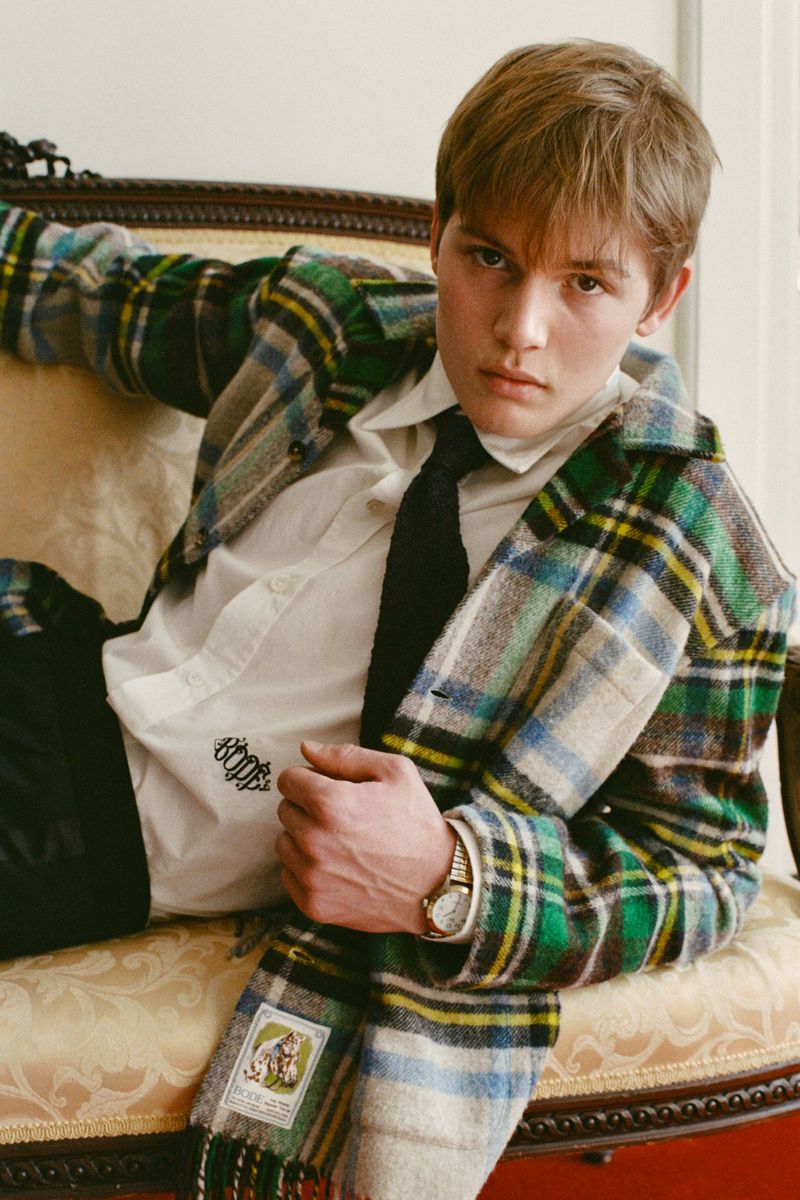 Young adult with blond hair wearing a plaid jacket and scarf, laying on an ornate chair. He has a wristwatch and a serious expression.