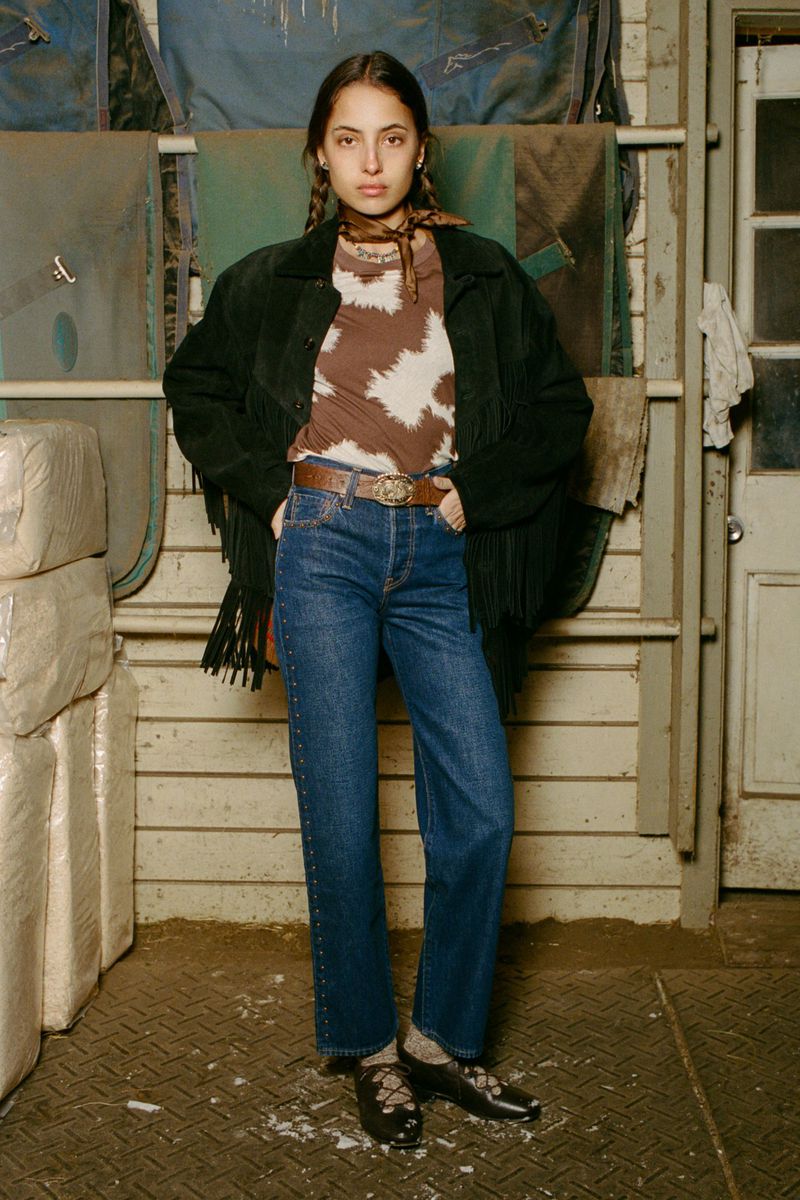 A woman poses indoors wearing a brown cow-print shirt, jeans, and a fringed black jacket. The background features blankets and a door.