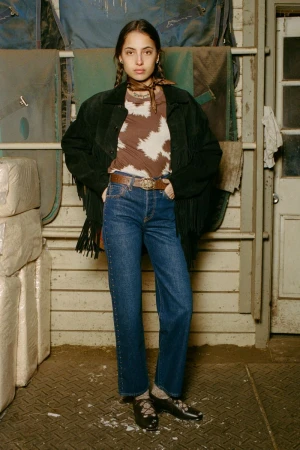 A woman poses indoors wearing a brown cow-print shirt, jeans, and a fringed black jacket. The background features blankets and a door.