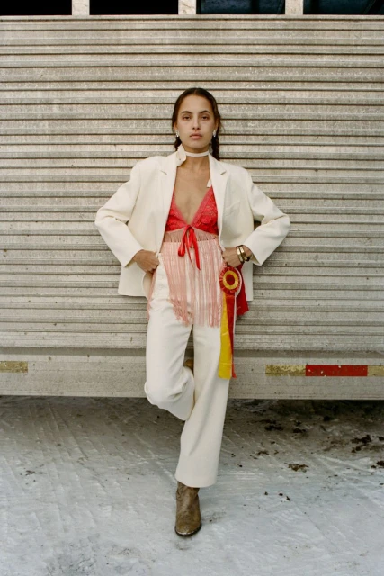 Stylish woman poses in a cream-colored suit, red top with fringe detail, and ankle boots against a textured metal wall outdoors.