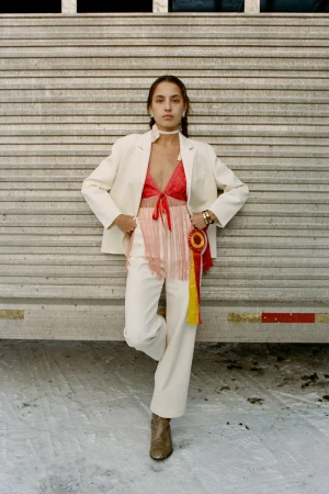 Stylish woman poses in a cream-colored suit, red top with fringe detail, and ankle boots against a textured metal wall outdoors.