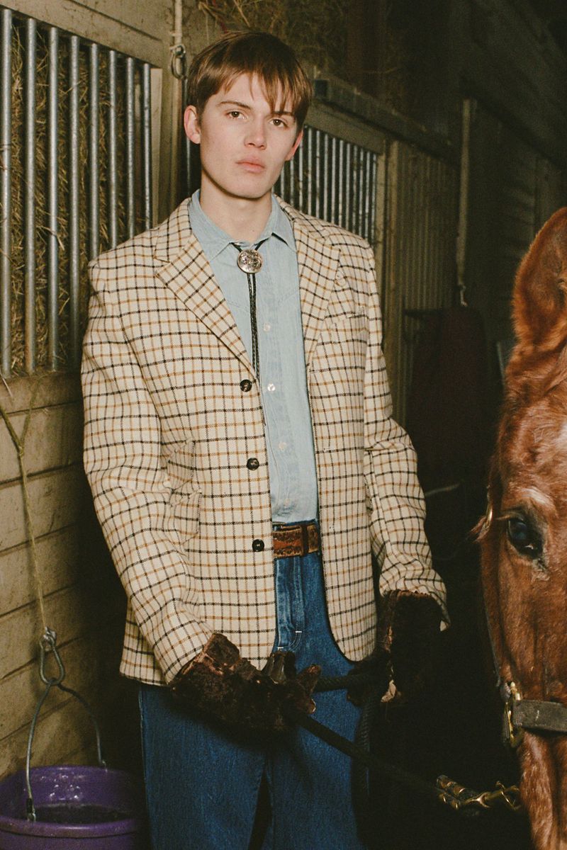 A young adult male wearing a plaid blazer poses with a horse inside of a barn with metal gates and dim lighting conditions.