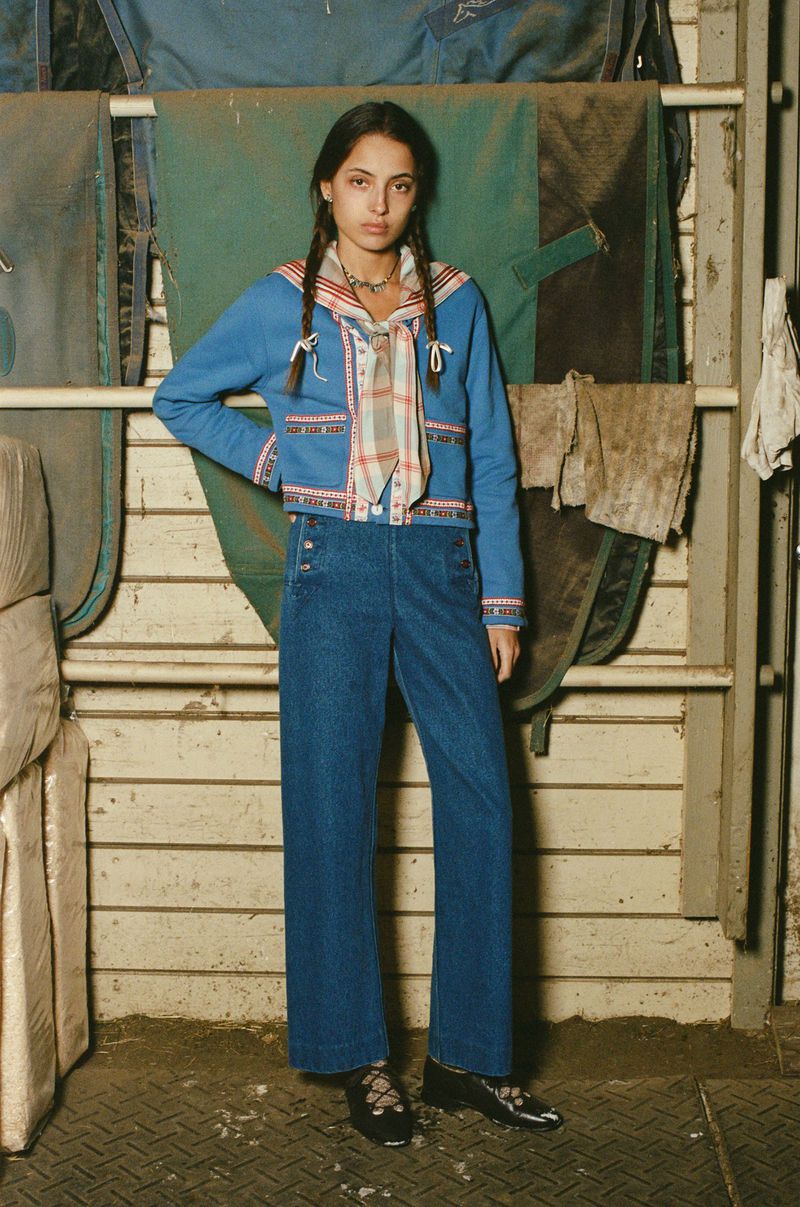 A young woman in blue denim wide-leg pants and a jacket with a scarf poses against a weathered wooden wall and horse blankets.