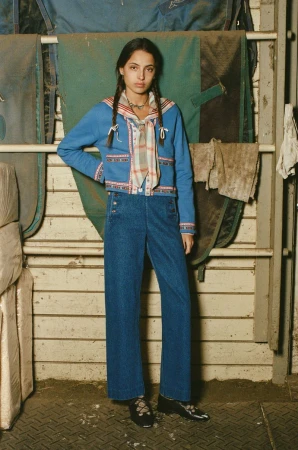 A young woman in blue denim wide-leg pants and a jacket with a scarf poses against a weathered wooden wall and horse blankets.