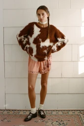 Model wearing a brown and white cow-pattern fuzzy sweater and patterned shorts standing against white siding.