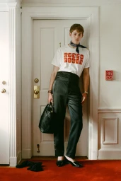 Model leaning against a white door wearing a graphic white t-shirt, dark trousers, and a dark scarf, holding a black handbag.