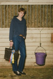 Model wearing a blue hoodie and jeans holding a studded handbag in a stable setting