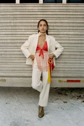 Model standing against a corrugated metal wall wearing a white blazer, white trousers, and a red fringed top with ribbons.