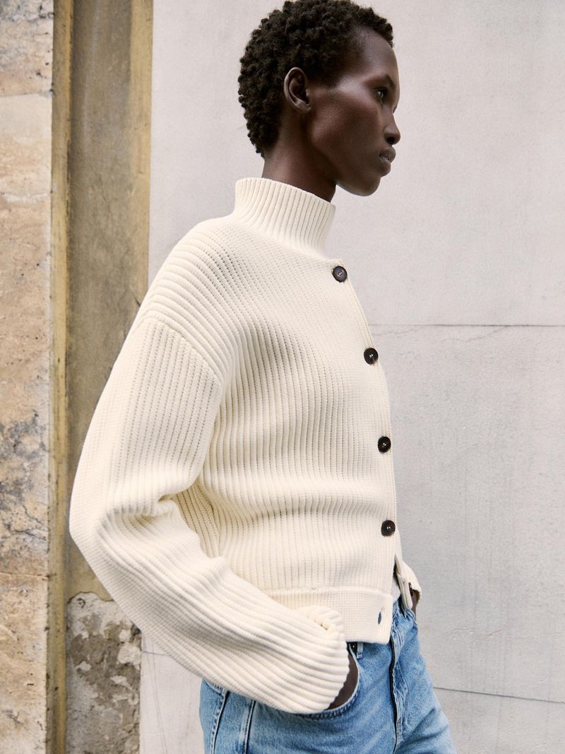 Adult woman poses in a cream-colored knit button-down cardigan and light blue jeans against a neutral wall in soft lighting.