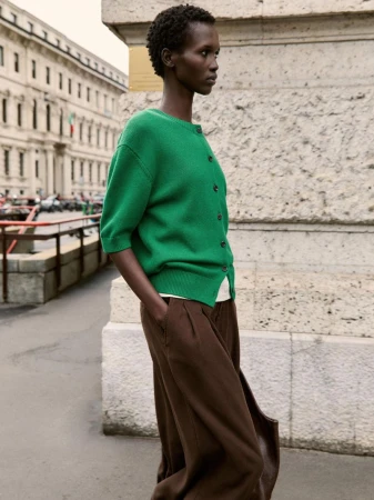 Woman in green sweater and brown pants poses in urban setting with neutral stone facade and building background.