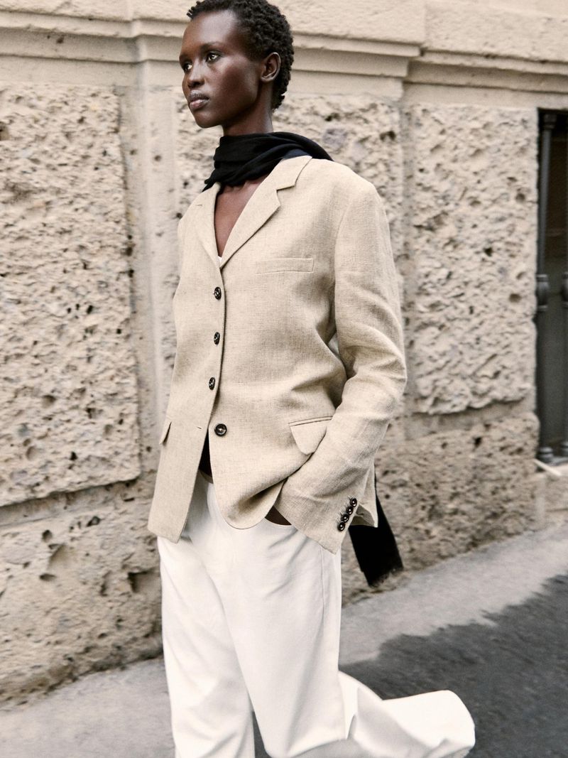 A woman in a beige blazer and white pants walks in front of a stone wall, showcasing a blend of fashion and urban style.