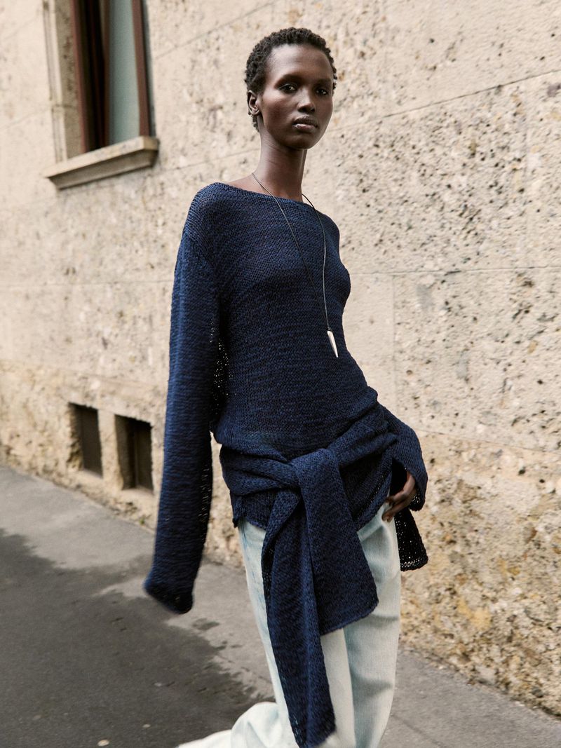 A slender African woman in a dark blue sweater and light blue jeans poses outside against a stone building on a city street.