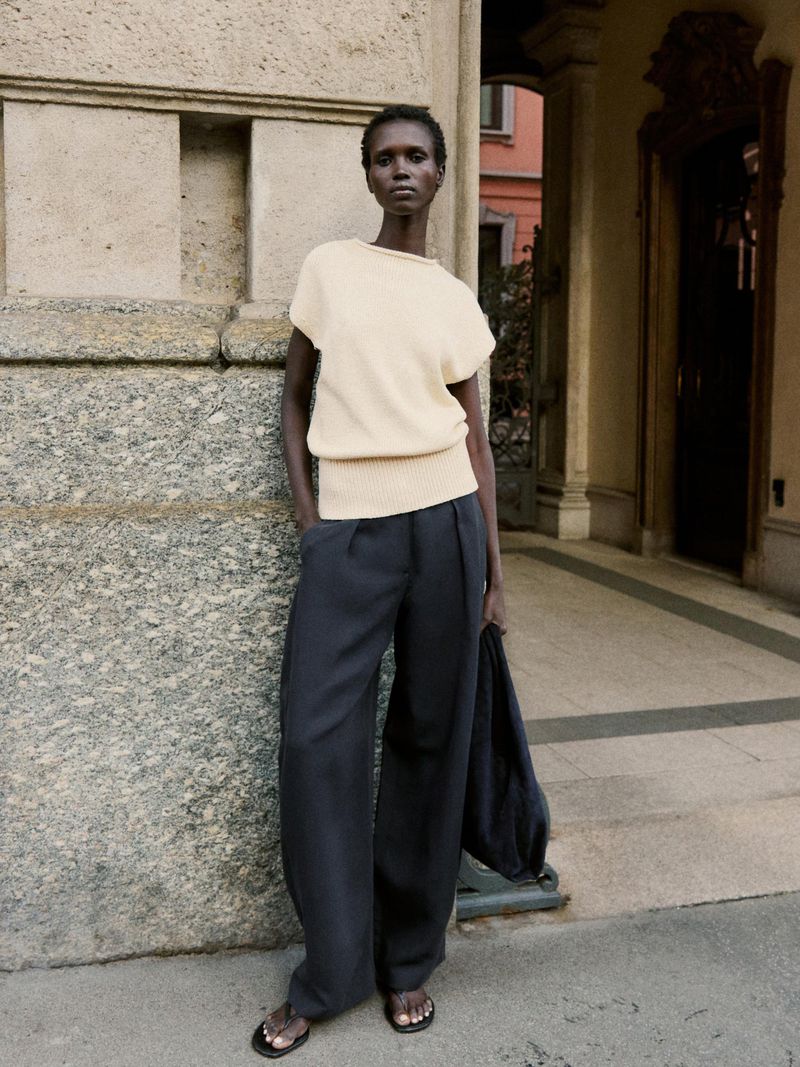 Fashionable woman in a cream-colored sweater and wide-leg pants leans against a stone wall in an urban environment, showcasing contemporary style.