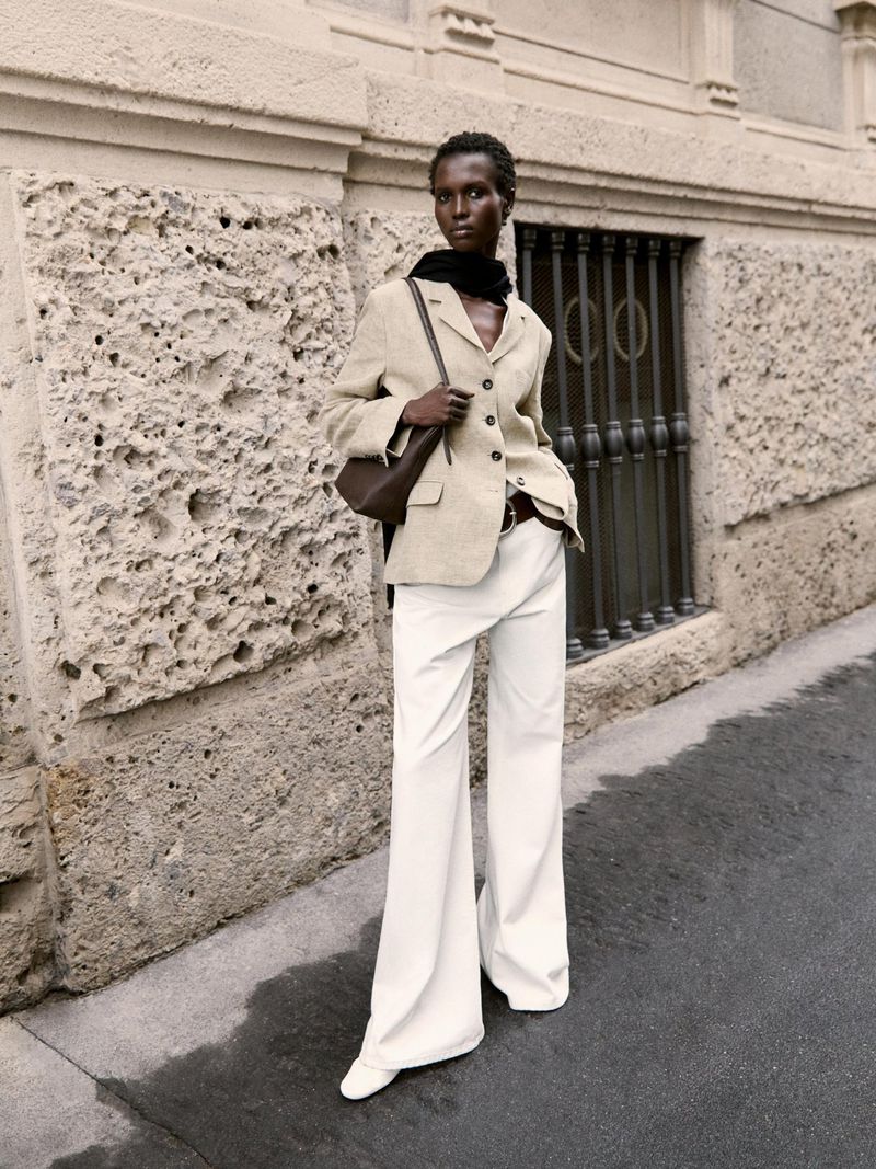 A stylish Black female model poses outdoors in beige blazer and white trousers with a brown handbag, in an urban setting.