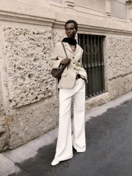 Amar Akway wearing a beige blazer and white trousers holding a brown leather handbag on a city street