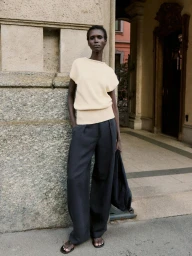 Model standing outdoors wearing a cream knit top and dark trousers next to a stone building column