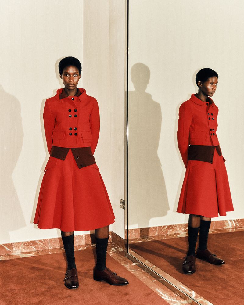 A black female fashion model is seen standing indoors in a red jacket and skirt, and black socks. She is also reflected in a mirror.