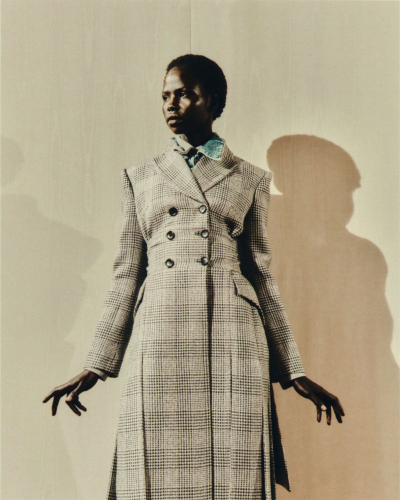 Elegant woman in a plaid coat, posing in a studio setting. The neutral backdrop highlights her outfit and silhouette, creating a fashion-forward image.