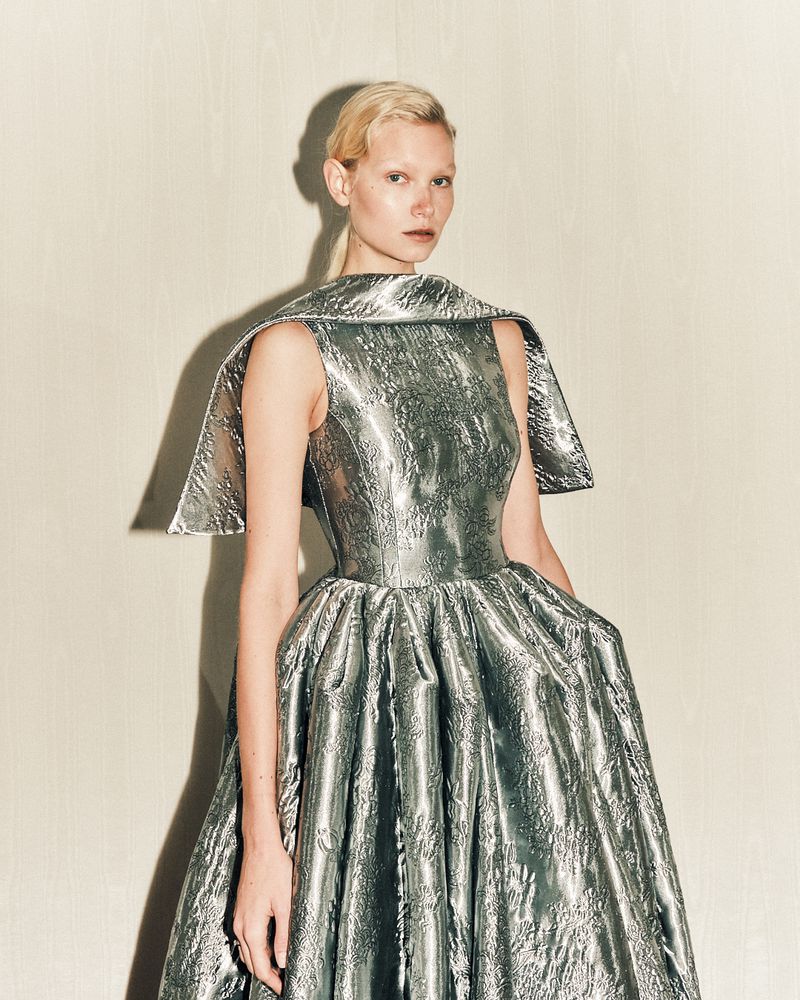 A woman with fair skin and blond hair is wearing a futuristic silver gown in a studio setting, against a neutral background.
