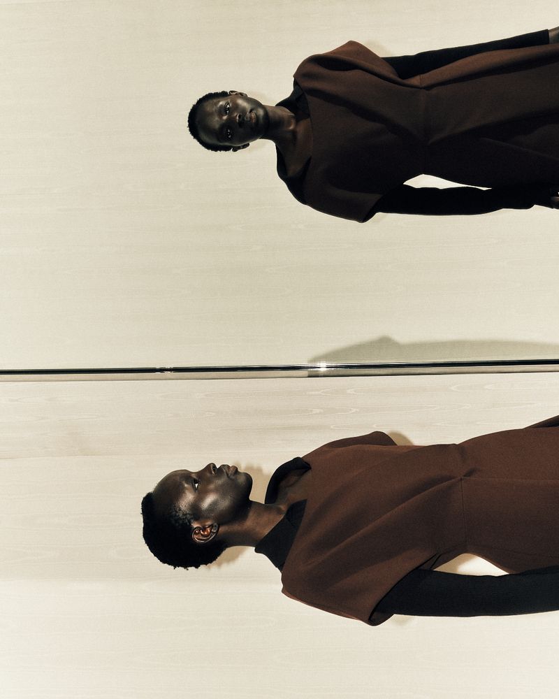 A fashion shot features a black female model in a brown dress and black top, reflected in a mirror with a neutral-toned background. 