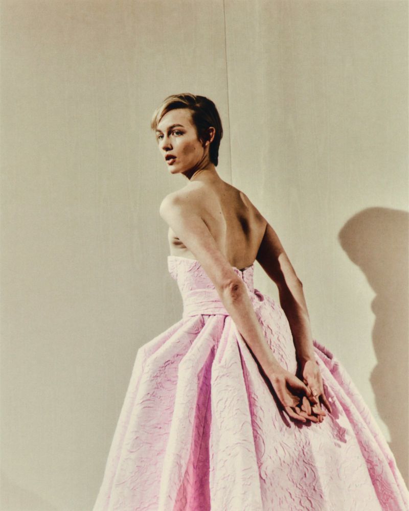 Elegant woman in a light pink strapless gown with hands clasped, gazing over her shoulder in a formal indoor setting with soft lighting.