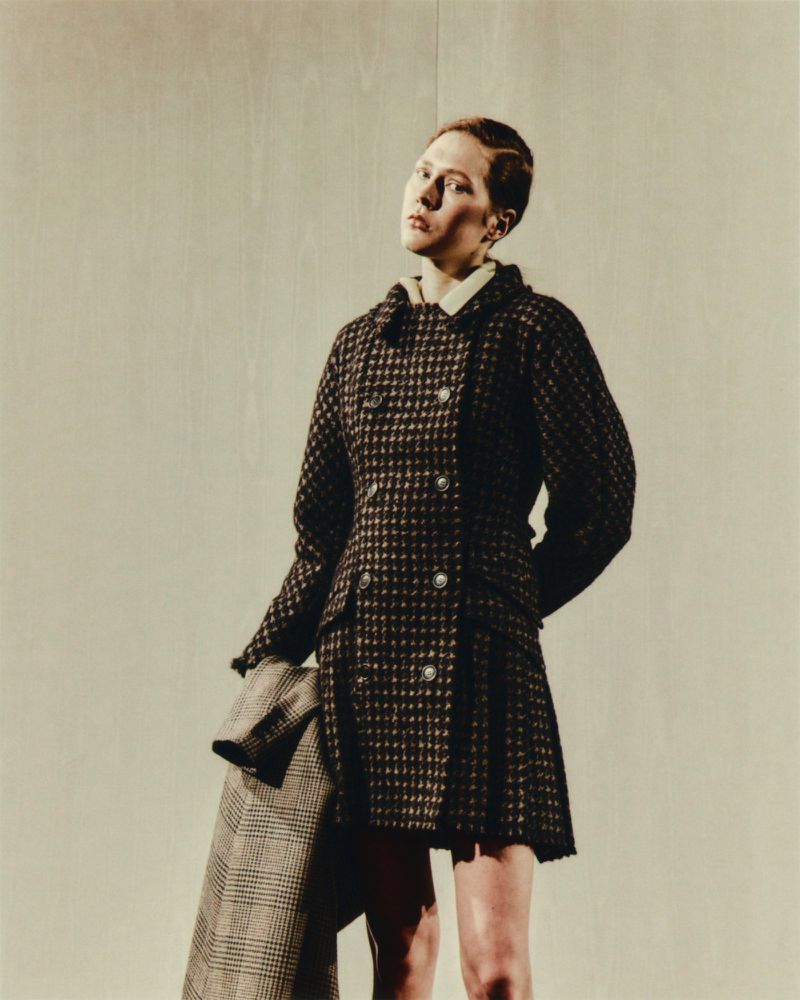 A woman poses in a brown and black houndstooth coat. She holds another coat of similar pattern. The background is an off-white wall.