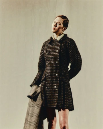 A woman poses in a brown and black houndstooth coat. She holds another coat of similar pattern. The background is an off-white wall.