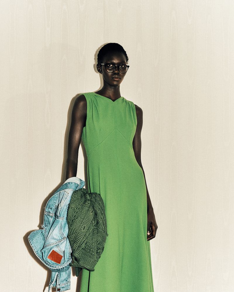 Dark-skinned female model in a green dress poses with a denim jacket and green knitted bag against a light beige backdrop.