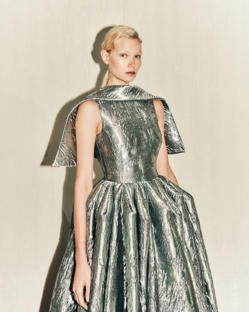 Model in a metallic silver structured evening gown posing against a light wood paneled wall.