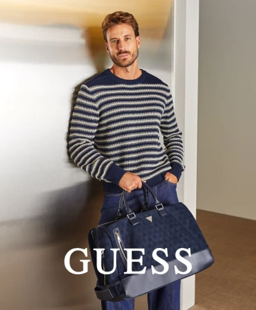 A man in a striped sweater and jeans leans against a wall, holding a navy travel bag with the Guess logo, showcasing a fashion shot.