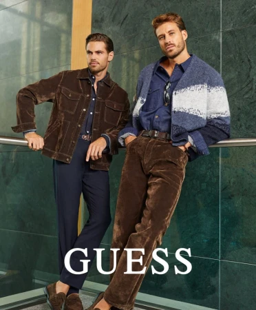 Two male fashion models pose in denim and corduroy jackets, accessorized with belts and loafers, in front of a marble tiled background.