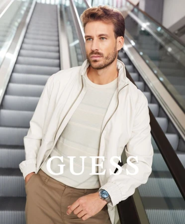 A man models a casual outfit consisting of a cream jacket, light striped tee shirt, and brown slacks, posed in front of an escalator.