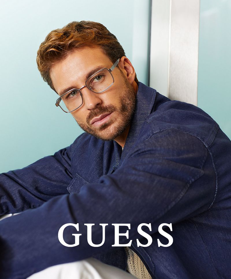 An adult male model with light brown hair, wearing eyeglasses and a blue denim jacket, poses against a light blue and silver background.