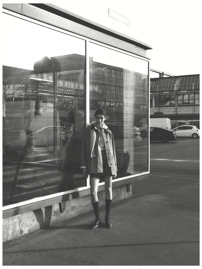 Monochrome portrait of a female fashion model posing in a coat and boots outdoors in an urban setting next to a window in daylight.