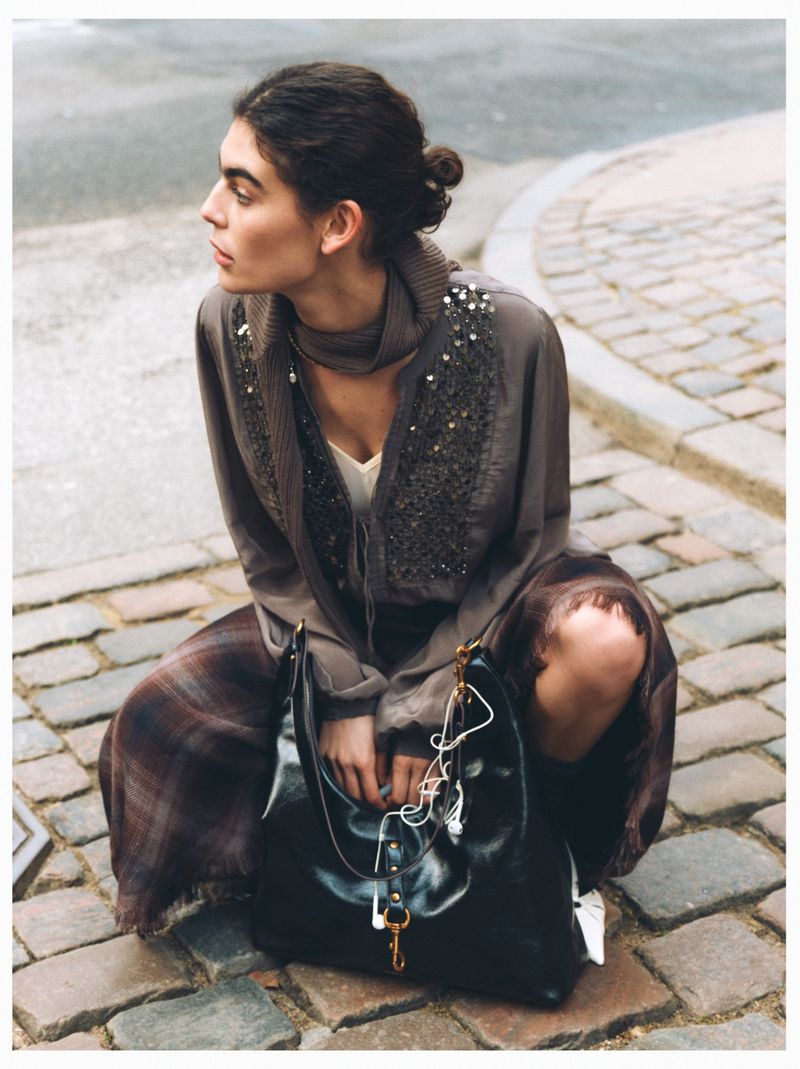 Adult female model poses in outdoor fashion shot with a brown beaded jacket, plaid skirt, and black leather purse on a cobblestone street.