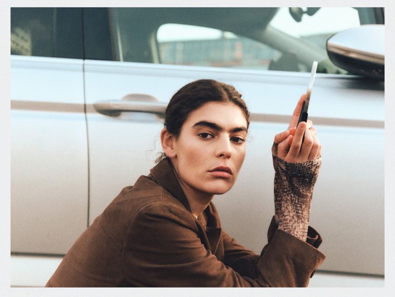Portrait of a woman in brown jacket seated in front of a silver SUV, holding a thin black object vertically, in an urban environment.