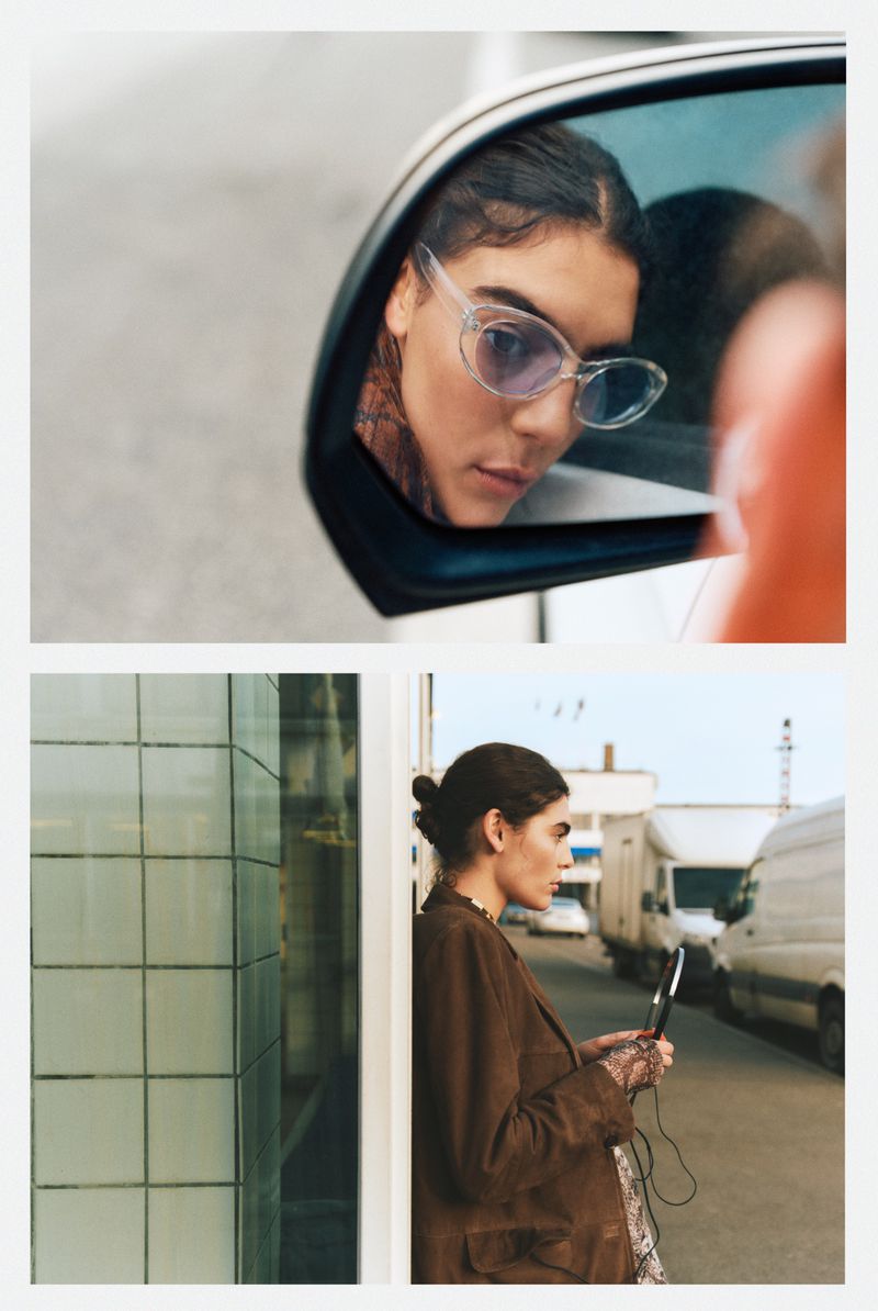 Woman reflected in car mirror wears glasses. Woman in brown blazer stands near tiled wall holding a phone on urban street.