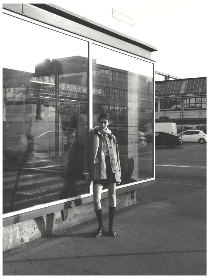 Model standing outdoors next to a large glass storefront window wearing a jacket and knee-high socks