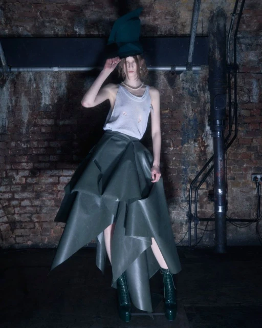 Fashionable young woman poses in an olive green high-low skirt, tank top, and matching hat and heels in a dark industrial setting.