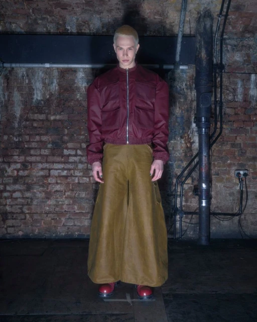 A person standing in an indoor space. The person wears a dark red jacket, light brown pants, and red shoes. Brick walls and pipes.