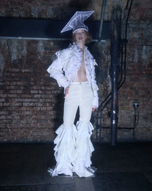 Adult female fashion model poses in front of a brick wall in a white ruffled outfit and headdress in a dimly lit indoor setting.