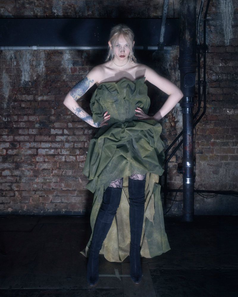 A fair-skinned adult woman poses in an olive dress and black boots against a brick wall backdrop with industrial pipe features.