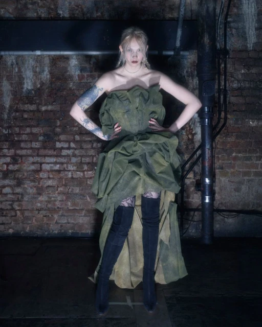 A fair-skinned adult woman poses in an olive dress and black boots against a brick wall backdrop with industrial pipe features.