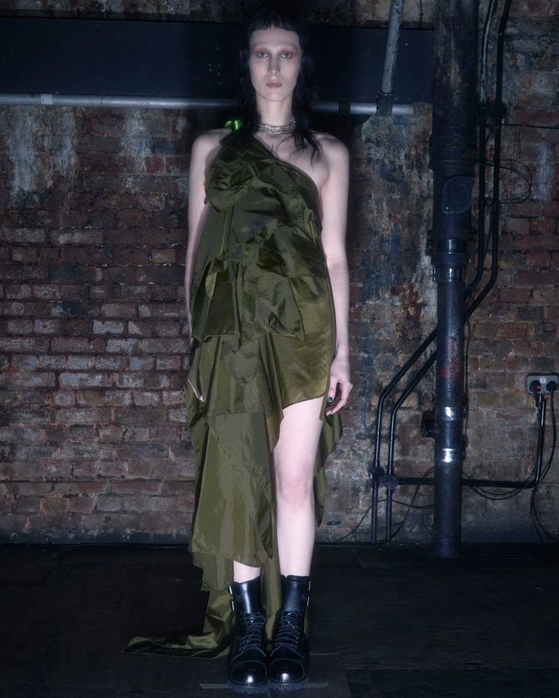 A slender adult female poses in an olive-green dress and black boots against a textured brick wall, illuminated by studio lighting.