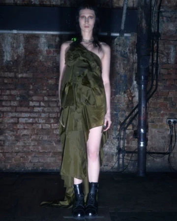 A slender adult female poses in an olive-green dress and black boots against a textured brick wall, illuminated by studio lighting.