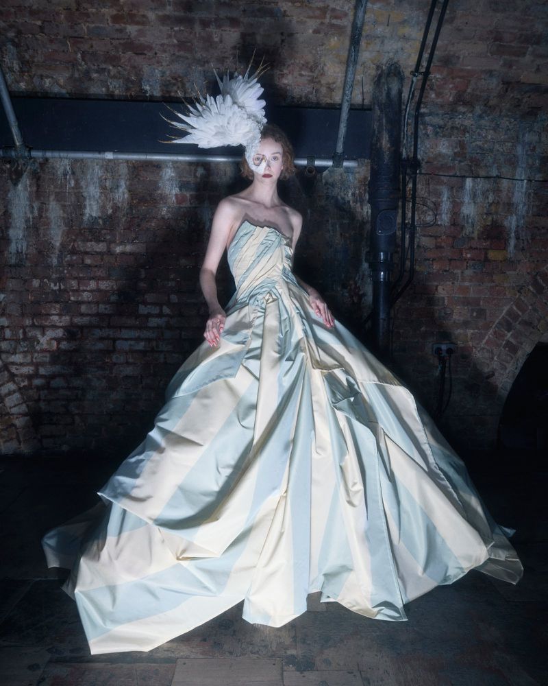 Woman in a striped strapless gown with a feathered headpiece stands in front of a brick wall with overhead piping in an underground space.