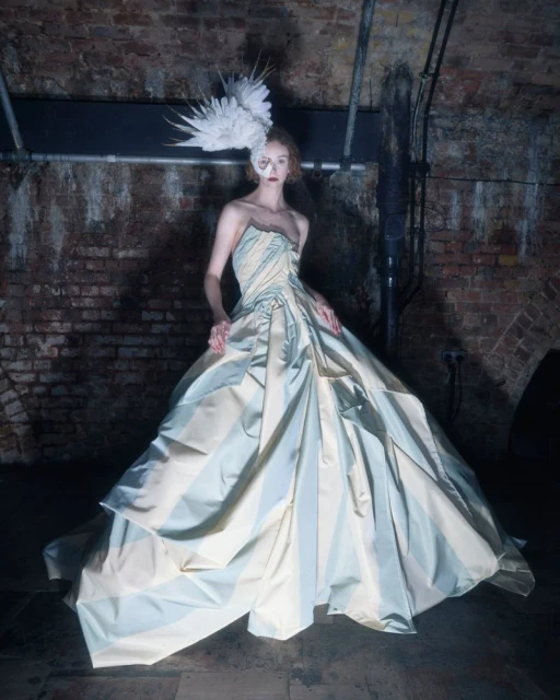 Woman in a striped strapless gown with a feathered headpiece stands in front of a brick wall with overhead piping in an underground space.