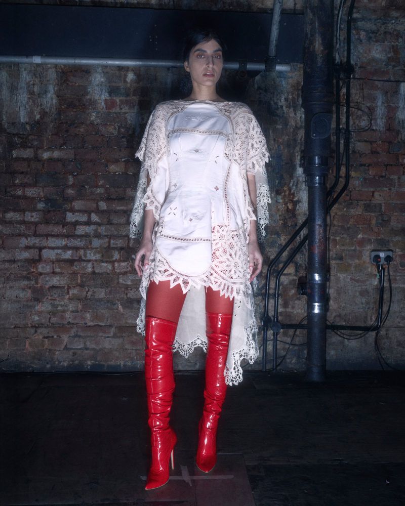 A woman stands in a white dress and red boots in an indoor setting, creating a fashion-forward and distinctive visual.