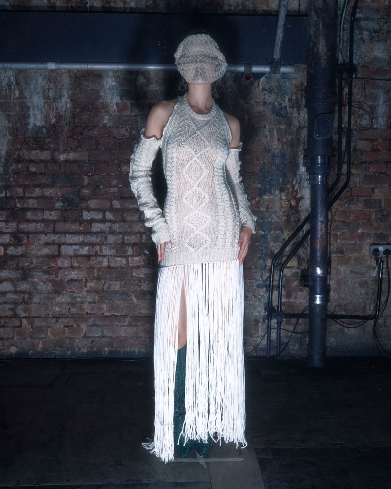 Woman in an off-white knit dress with a fringe skirt, wearing a matching balaclava, standing in a dimly lit brick-walled room.