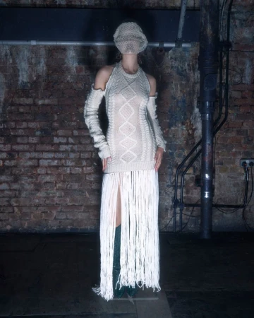 Woman in an off-white knit dress with a fringe skirt, wearing a matching balaclava, standing in a dimly lit brick-walled room.
